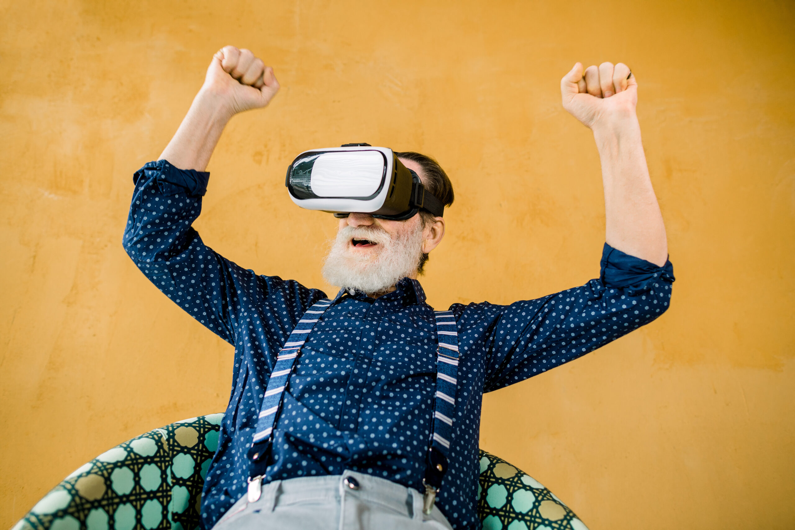 Excited Joyful Senior Bearded Man In Stylish Dark Blue Shirt And Suspenders, Using Vr 3d Glasses And Watching Film Or Football Game With Smile And Clenched Fists. Studio Shot On Yellow Background