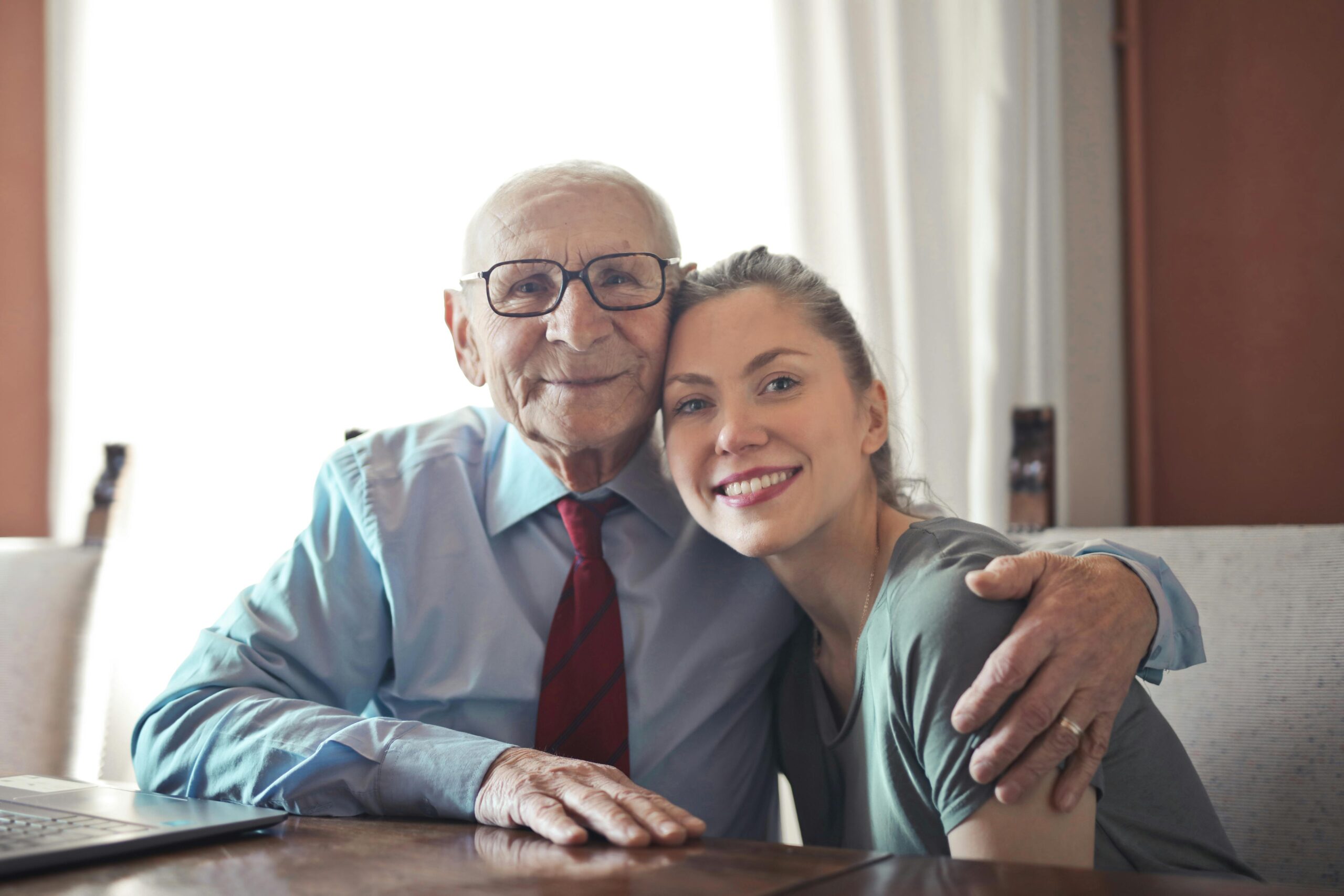 positive-senior-man-in-formal-wear-and-eyeglasses-hugging-with-young-lady-while-sitting-at-table